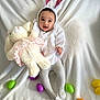 baby, bunny_ears, plush_toy, stuffed_animal, easter_eggs, white_blanket, tights, smiling, cute, child, holiday, costume, infant, indoors, celebration, soft_toy, happy, playful, festive, portrait