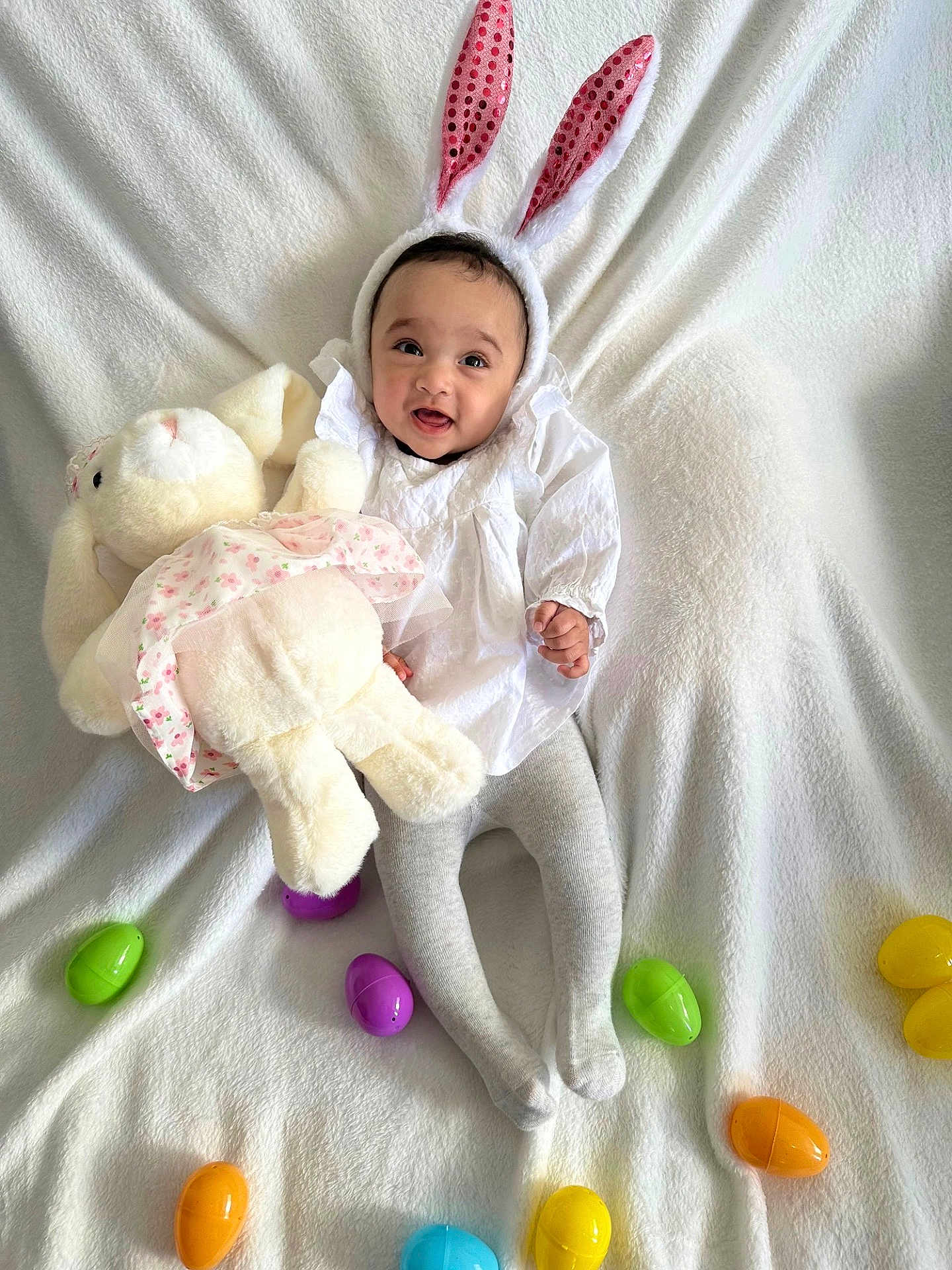 Noeli is registered to the contest to win money with this photo: baby, bunny_ears, plush_toy, stuffed_animal, easter_eggs, white_blanket, tights, smiling, cute, child, holiday, costume, infant, indoors, celebration, soft_toy, happy, playful, festive, portrait