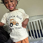 Ezra joined the competition — help win amazing prizes! toddler, child, smiling, curly_hair, tshirt, tractor, text_on_clothing, pants, crib, bedroom, bedding, animal_print, happy, indoors, furniture, person, young_child, cute, playful, home