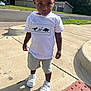 Ezra is registered to the contest to win money with this photo: toddler, child, sidewalk, street, dinosaur_tshirt, shorts, white_sneakers, shadow, outdoor, daylight, curly_hair, urban, concrete, traffic_sign, grass, building, greenery, person, casual_clothing, summer