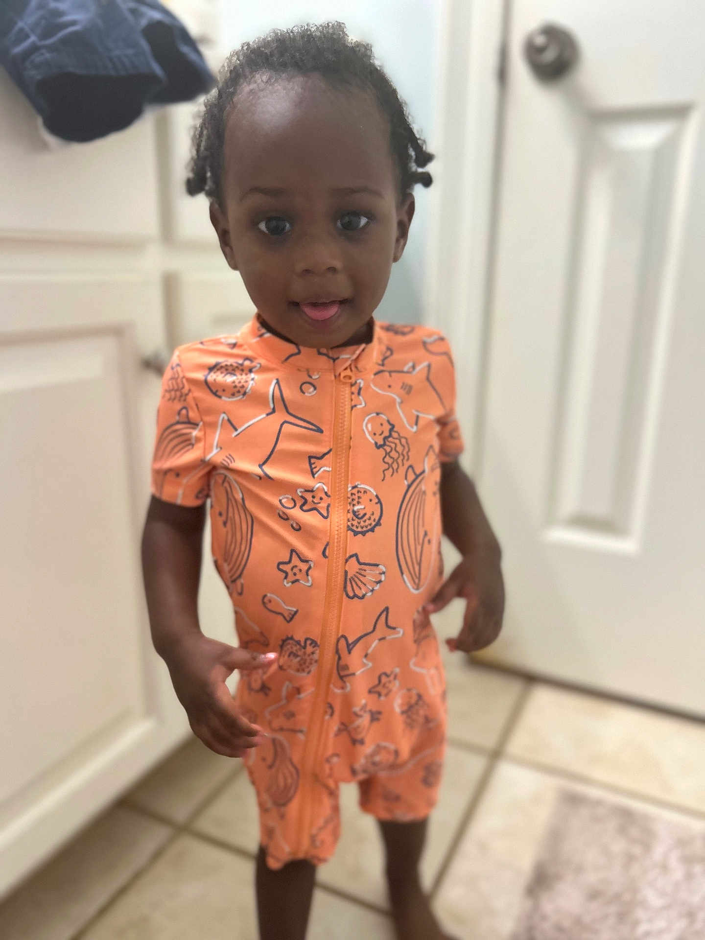Ezra is registered to the contest to win money with this photo: toddler, child, indoors, orange_clothing, onesie, standing, barefoot, bathroom, tile_floor, door, cabinet, curly_hair, playful, tongue_out, young_child, person, casual, home, cute, curious