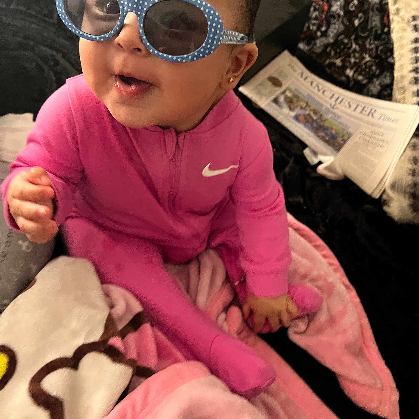 Sarah joined the competition — help win amazing prizes! baby, blanket, child, cozy, cute, face, floor, happy, head, indoor, newspaper, nike_logo, person, pink_clothing, polka_dots, portrait, seated, smiling, sunglasses, toy