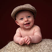 Gunner is registered to the contest to win money with this photo: baby, background, blue_eyes, cap, child, chubby_arms, closeup, cute, expression, face, happy, hat, infant, knitted_blanket, portrait, resting, skin, smiling, studio_photo, warm_colors