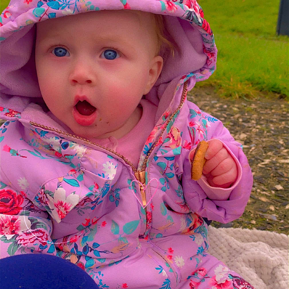 Orla is registered to the contest to win money with this photo: baby, bonnet, clothing, coat, face, grass, hat, head, hood, hoodie, jacket, knitwear, outdoors, person, photography, plant, portrait, purple, sweater, sweatshirt