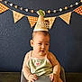 baby, birthday_hat, high_chair, toddler, face, child, indoor, celebration, decorations, banner, pom_poms, blue_wall, orange, green_clothing, cute, young_child, portrait, smiling, sitting, party