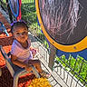 child, toddler, girl, sitting, playground, outdoor, chalkboard, chalk_drawing, chair, fence, sunlight, purple_clothing, hair_bows, sandals, carpet, portrait, face, looking_at_camera, play_area, colorful