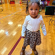 Hannah is registered to the contest to win money with this photo: child, toddler, portrait, standing, gymnasium, indoor, wooden_floor, basketball_court, leopard_print, skirt, white_shirt, boots, necklace, hair_clips, braids, fashion, serious_expression, colorful_decorations, balloons, speakers