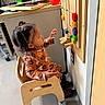 child, toddler, girl, indoor, playroom, wooden_chair, activity_panel, educational_toy, colorful, reaching, curious, profile, braided_hair, floral_clothing, sitting, furniture, wall, cabinet, home, photograph