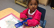 Hannah is registered to the contest to win money with this photo: child, toddler, girl, portrait, sitting, classroom, art, painting, watercolor, paintbrush, palette, table, chair, creative, focused, looking_at_camera, pink_clothing, braided_hair, indoor, school