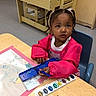child, toddler, girl, portrait, sitting, classroom, art, painting, watercolor, paintbrush, palette, table, chair, creative, focused, looking_at_camera, pink_clothing, braided_hair, indoor, school