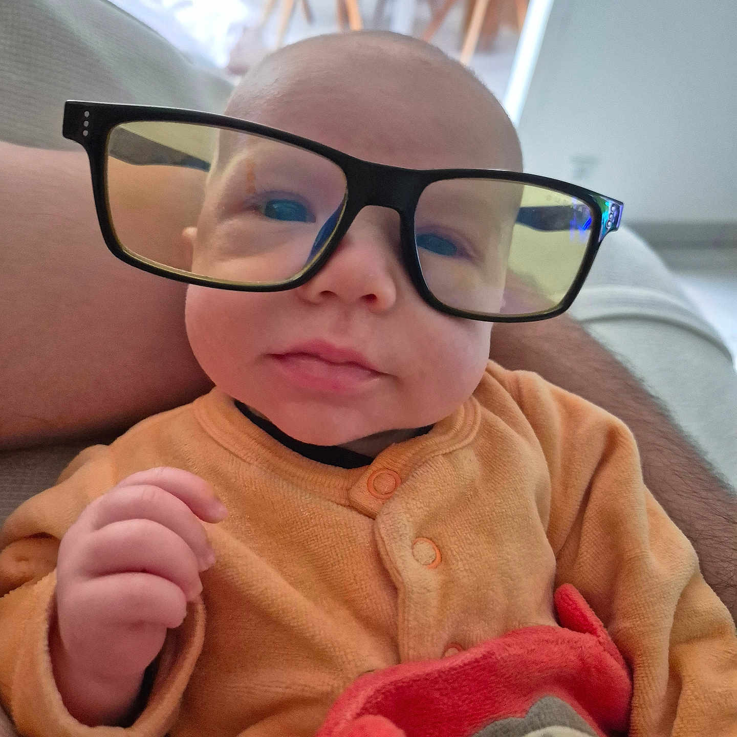 Maël a rejoint le concours — aidez-le/la à gagner de superbes lots ! baby, chair, child, cozy, cute, face, floor, furniture, glasses, hand, head, indoor, infant, living_room, orange_onesie, person, plush_toy, portrait, sitting, soft_light