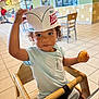 Aniyah is registered to the contest to win money with this photo: toddler, child, high_chair, donut, food, hat, paper_hat, curly_hair, earrings, blue_shirt, shorts, indoor, restaurant, krispy_kreme, table, chair, tile_floor, person, holding, sitting