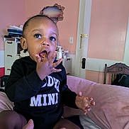 Daron joined the competition — help win amazing prizes! toddler, child, indoor, hand, food, face, clothing, black_outfit, fingers, mouth, furniture, door, pink_wall, cabinet, curious, feeding, person, sitting, expression, room