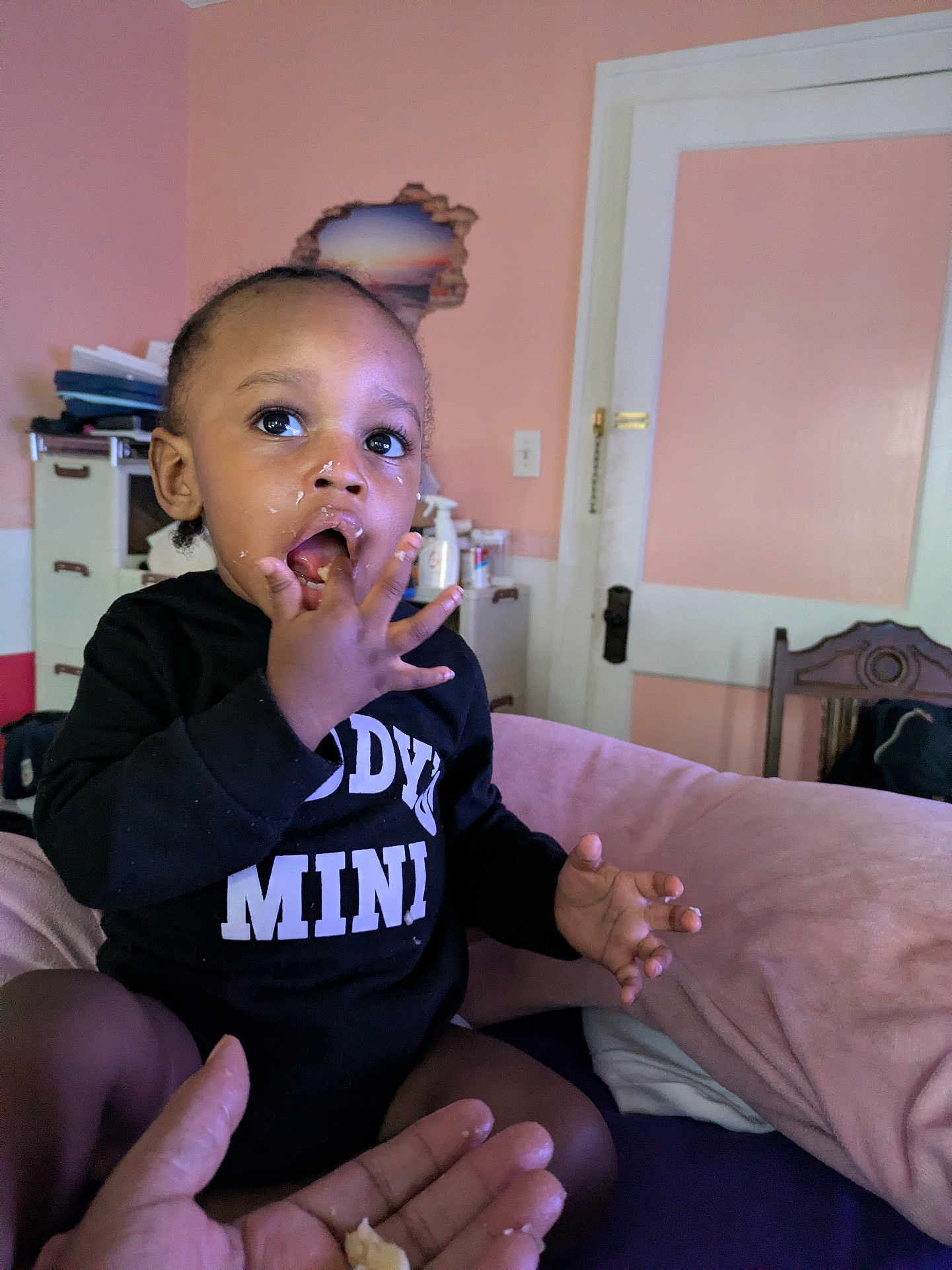 Daron joined the competition — help win amazing prizes! toddler, child, indoor, hand, food, face, clothing, black_outfit, fingers, mouth, furniture, door, pink_wall, cabinet, curious, feeding, person, sitting, expression, room