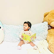 Nia joined the competition — help win amazing prizes! toddler, child, book, reading, plush_toy, stuffed_animal, bed, pillow, cushion, indoor, curly_hair, headband, socks, cozy, soft, cute, playful, happy, light, portrait