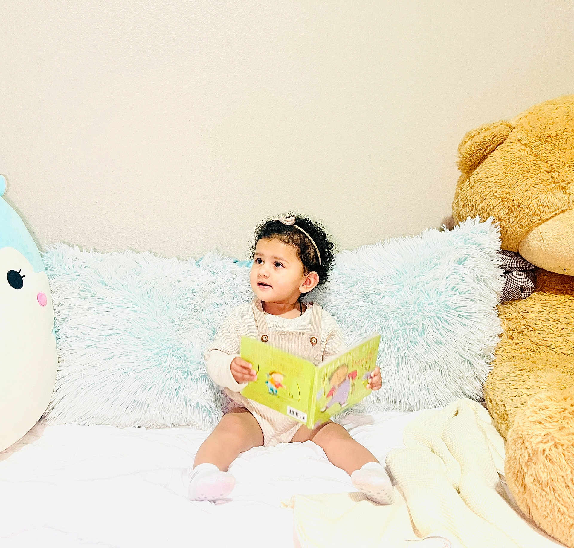 Nia joined the competition — help win amazing prizes! toddler, child, book, reading, plush_toy, stuffed_animal, bed, pillow, cushion, indoor, curly_hair, headband, socks, cozy, soft, cute, playful, happy, light, portrait