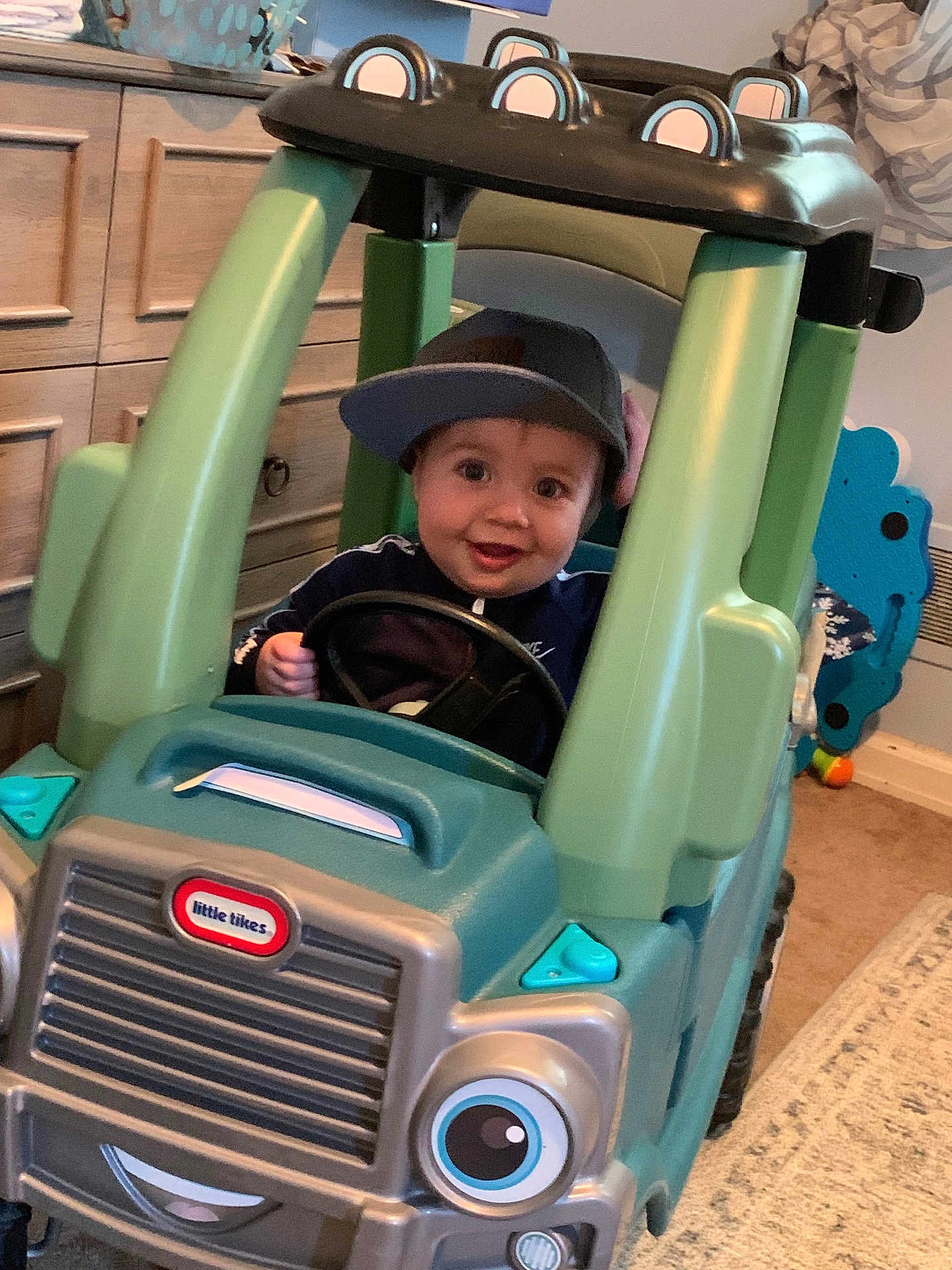 Declan is registered to the contest to win money with this photo: toddler, child, toy_car, little_tikes, cap, indoor, play, smile, face, steering_wheel, car, plastic, furniture, carpet, drawer, happy, hat, room, child_playing, fun