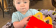 Declan is registered to the contest to win money with this photo: toddler, child, happy_meal_box, batman_blanket, restaurant, high_chair, curly_hair, indoor, table, chair, floor, person, clothing, smile, food_container, modern_interior, light, seat, baby, cute