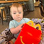 toddler, child, happy_meal_box, batman_blanket, restaurant, high_chair, curly_hair, indoor, table, chair, floor, person, clothing, smile, food_container, modern_interior, light, seat, baby, cute