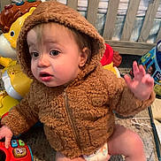 Declan is registered to the contest to win money with this photo: baby, child, hoodie, brown_clothing, toy, carpet, indoor, diaper, furniture, crib, colorful, plastic_toy, curious, sitting, hand_raised, expression, face, young_child, playtime, room
