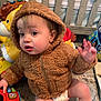baby, child, hoodie, brown_clothing, toy, carpet, indoor, diaper, furniture, crib, colorful, plastic_toy, curious, sitting, hand_raised, expression, face, young_child, playtime, room