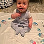baby, child, smiling, sitting, onesie, play_mat, indoor, floor, crib, pillow, dinosaur_pattern, happy, cute, infant, clothing, hand, face, eyes, toy, carpet
