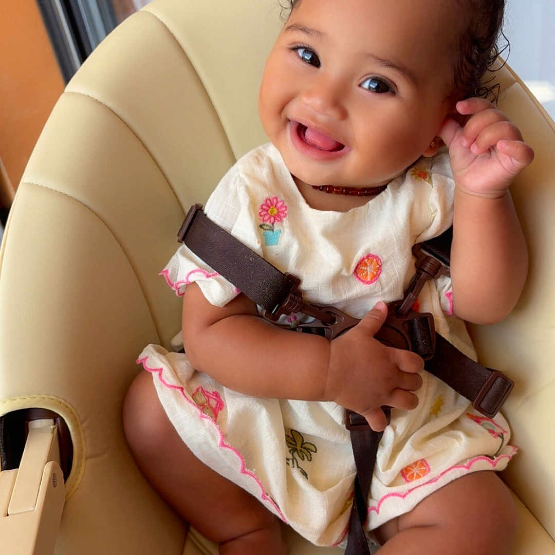 Syarah participe au concours pour gagner de l'argent avec cette photo : baby, child, cute, dress, embroidery, face, foot, furniture, hand, happy, high_chair, indoors, person, pink_bows, playful, seatbelt, skin, smiling, toddler, window