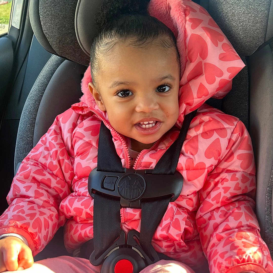 Xoe is registered to the contest to win money with this photo: child, toddler, car_seat, pink_jacket, heart_pattern, safety_harness, smiling, happy, indoors, seat_belt, car, sunlight, young_child, cute, portrait, clothing, hair_puff, seat, baby, casual
