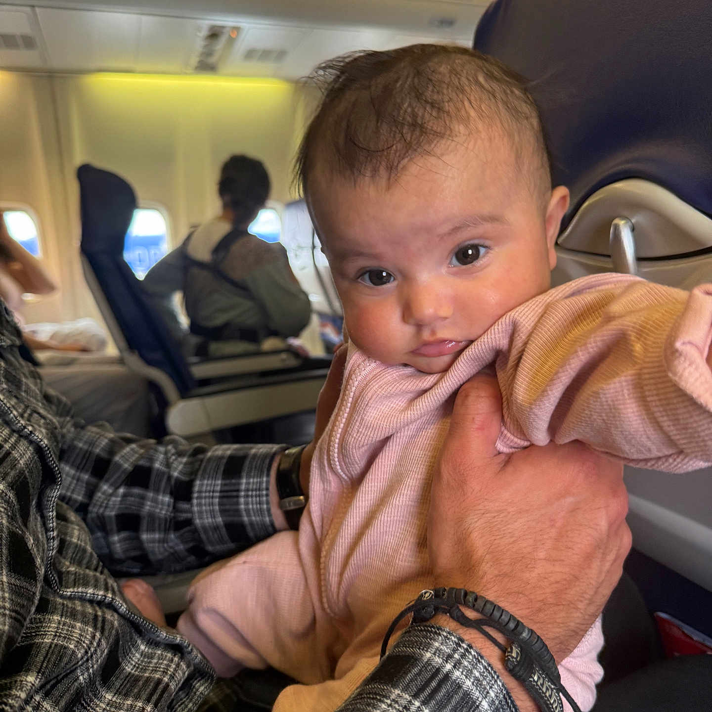 Demi joined the competition — help win amazing prizes! adult_hand, airplane_cabin, baby, bracelet, child, clothing, face, holding, infant, interior, looking, passenger, person, pink_onesie, plaid_shirt, portrait, seat, seatback, travel, window