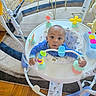 Mason is registered to the contest to win money with this photo: baby, child, infant, activity_center, jumper, toy, playroom, indoor, sitting, curious, face, eyes, blue_clothing, colorful, plush_toy, buttons, rug, floor, furniture, portrait