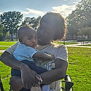 Mason is registered to the contest to win money with this photo: baby, infant, child, adult, parent, 抱抱, portrait, outdoor, park, stroller, family, holding, sitting, sunlight, backlighting, grass, trees, bench, blue_shirt, striped_sock