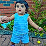 toddler, child, person, blue_clothing, shorts, crocheted_top, black_shoes, purple_socks, hand_holding, brick_wall, plants, outdoor, pavement, walking, cute, young_child, expression, greenery, summer, daylight