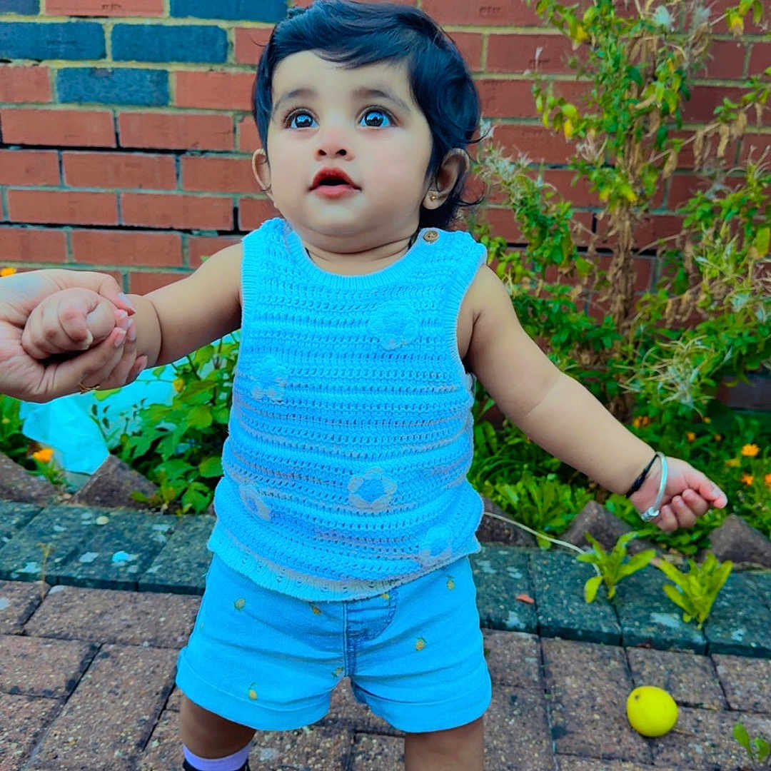 Avinaaz is registered to the contest to win money with this photo: black_shoes, blue_clothing, brick_wall, child, crocheted_top, cute, daylight, expression, greenery, hand_holding, outdoor, pavement, person, plants, purple_socks, shorts, summer, toddler, walking, young_child