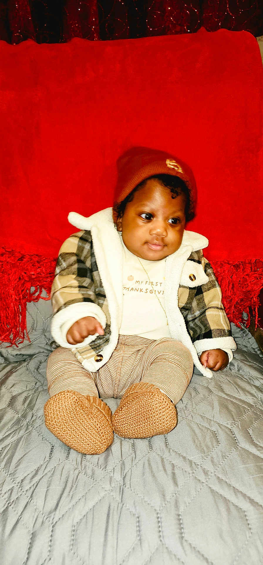 Joriel is registered to the contest to win money with this photo: baby, child, infant, beanie, plaid_jacket, knitted_booties, blanket, quilt, red_background, cute, cozy, fall_clothing, sitting, indoors, portrait, warm_colors, curly_hair, soft_expression, casual, comfortable