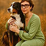 affection, animal, bernese_mountain_dog, brown_hair, dog, earrings, fall, friendship, glasses, golden_hour, grass, green_dress, hug, kneeling, nature, outdoor, pet, portrait, warm_lighting, woman