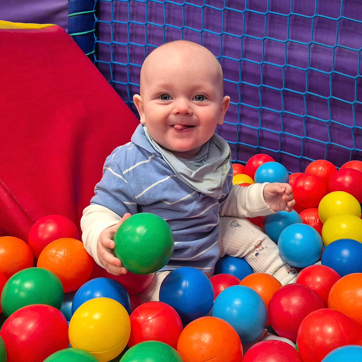 Grayson is registered to the contest to win money with this photo: baby, ball_pit, blue_shirt, child, colorful_balls, cute, fun, happy, indoor, infant, multicolor, netting, padded_surface, person, play_area, playful, sitting, smiling, tongue_out, toy