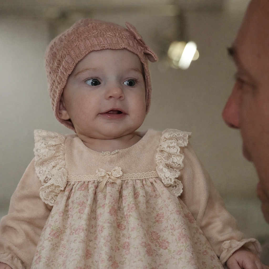 Elizabeth a rejoint le concours — aidez-le/la à gagner de superbes lots ! baby, child, infant, dress, knitted_hat, bow, person, face, portrait, curious, cute, soft_lighting, indoor, adult, interaction, closeup, expression, warm, family, clothing