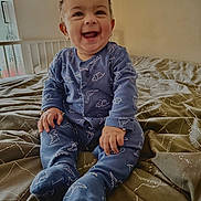 Aveer joined the competition — help win amazing prizes! baby, child, smiling, blue_onesie, dinosaur_pattern, bed, bedspread, indoor, happy, sitting, person, face, cute, infant, portrait, natural_light, cozy, hands, feet, room