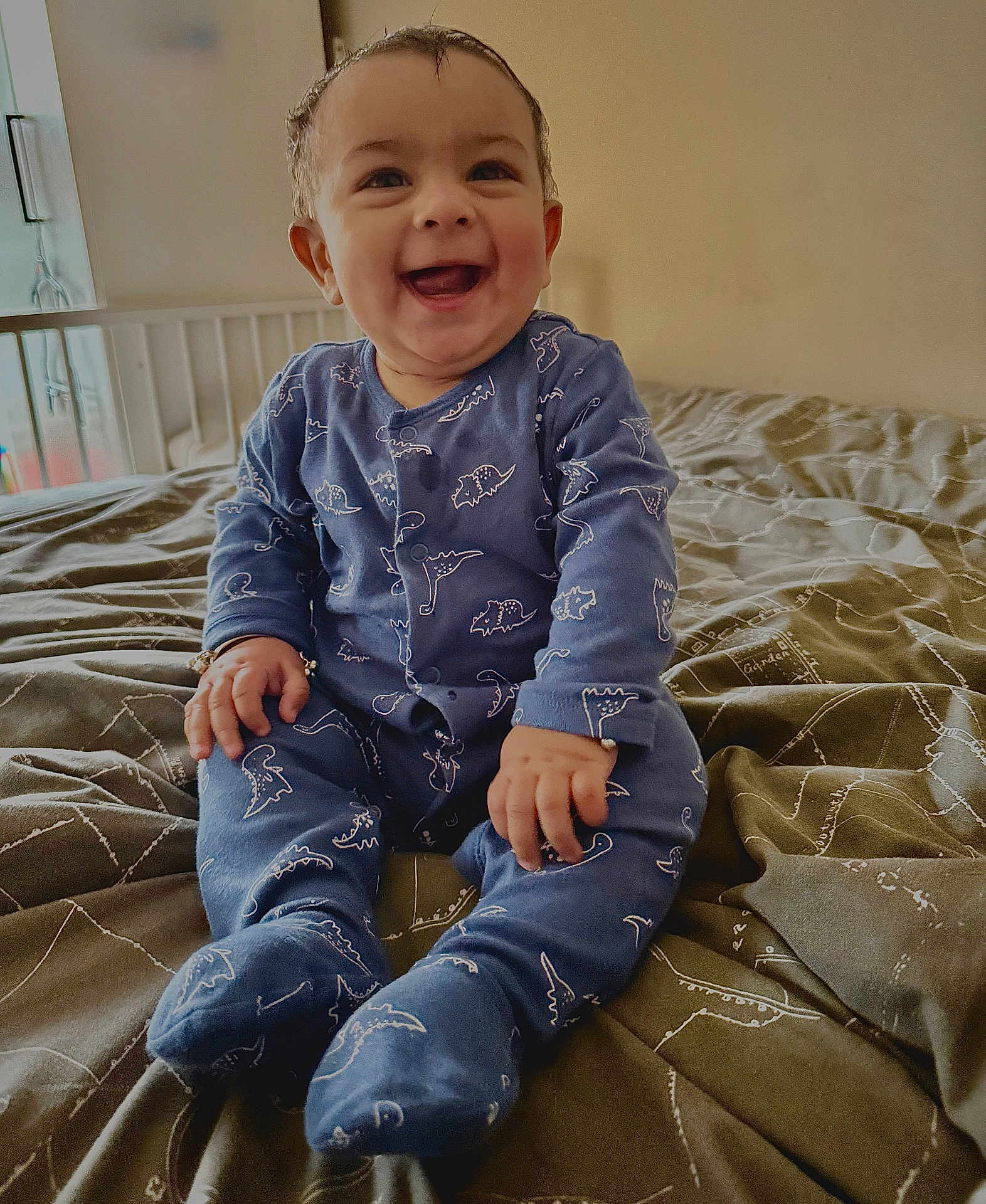 Aveer joined the competition — help win amazing prizes! baby, child, smiling, blue_onesie, dinosaur_pattern, bed, bedspread, indoor, happy, sitting, person, face, cute, infant, portrait, natural_light, cozy, hands, feet, room