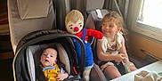 Liam participe au concours pour gagner de l'argent avec cette photo : baby, car_seat, child, clothing, curly_hair, cute, happy, indoor, knitted_doll, person, playful, seat, seatbelt, smile, sunlight, toy, vehicle, white_dress, window, yellow_overalls