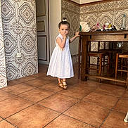 Alia participe au concours pour gagner de l'argent avec cette photo : child, girl, white_dress, sandals, indoor, kitchen, wooden_table, tiles, floor, furniture, curious, person, standing, hand_on_table, decor, bottle, bag, hair_tie, expression, young