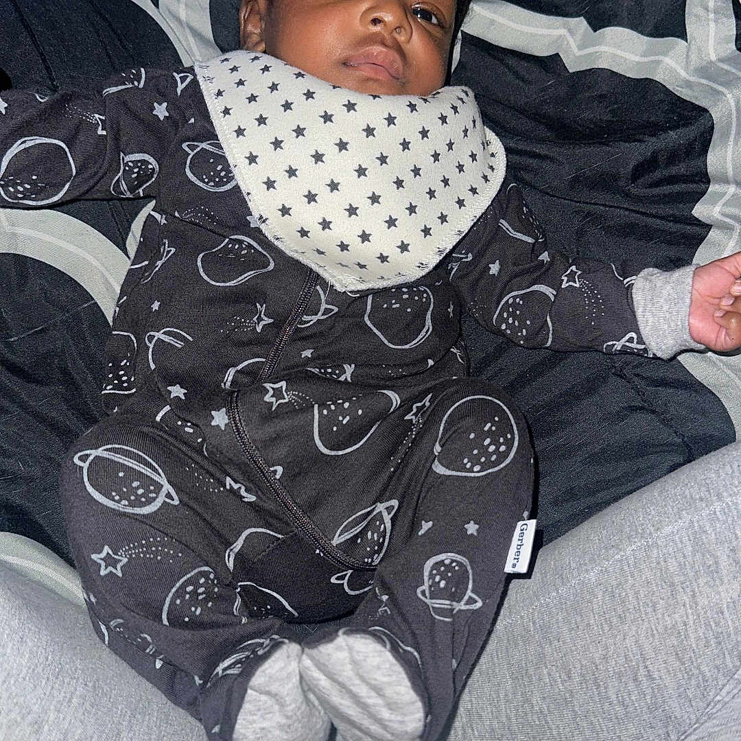 Jeremiah joined the competition — help win amazing prizes! baby, infant, onesie, bib, space_theme, stars, blanket, lying_down, indoor, cute, calm, child, person, sleepwear, cozy, soft_texture, black_clothing, patterned_clothing, headwear, portrait