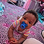 Isiah is registered to the contest to win money with this photo: baby, pacifier, blanket, pattern, crown, striped_clothing, infant, bedroom, toy, storage_bin, clothing, furniture, child, indoors, soft_toy, pink, black, purple, curly_hair, hands