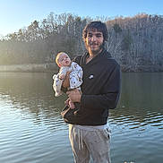 Weston joined the competition — help win amazing prizes! baby, man, lake, dock, water, trees, sunset, outdoor, nature, blanket, holding, casual_clothing, smile, beard, hoodie, pants, calm, reflection, wood, person