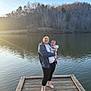 Weston is registered to the contest to win money with this photo: woman, baby, dock, lake, water, barefoot, trees, sunlight, smile, holding, outdoor, nature, wood, blanket, person, calm, sky, reflection, casual_clothing, relaxed