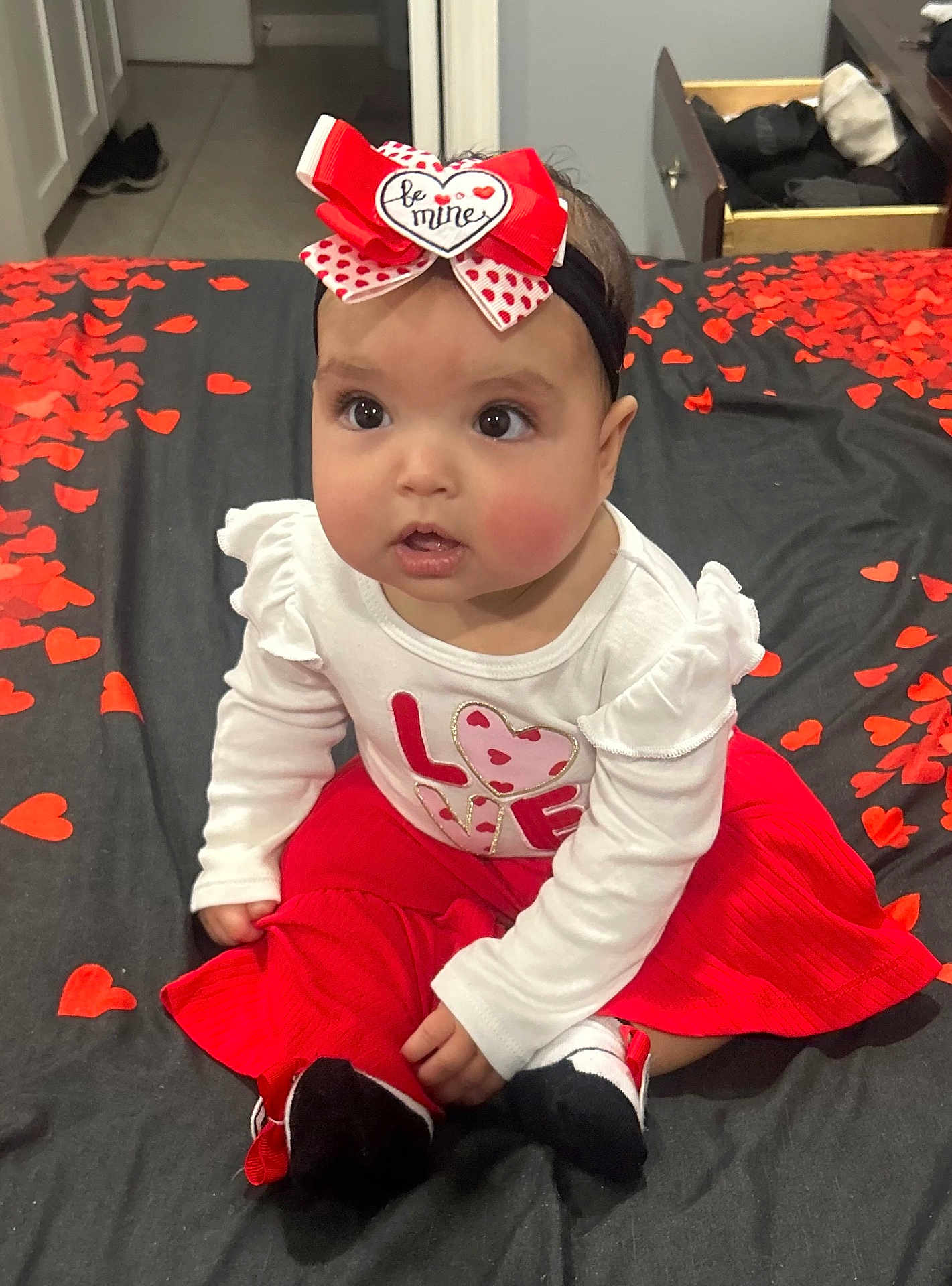 Marlenys is registered to the contest to win money with this photo: baby, child, girl, headband, heart, love, bed, bedroom, red_skirt, white_top, sitting, indoor, cute, petals, decoration, clothing, footwear, expression, portrait, innocence