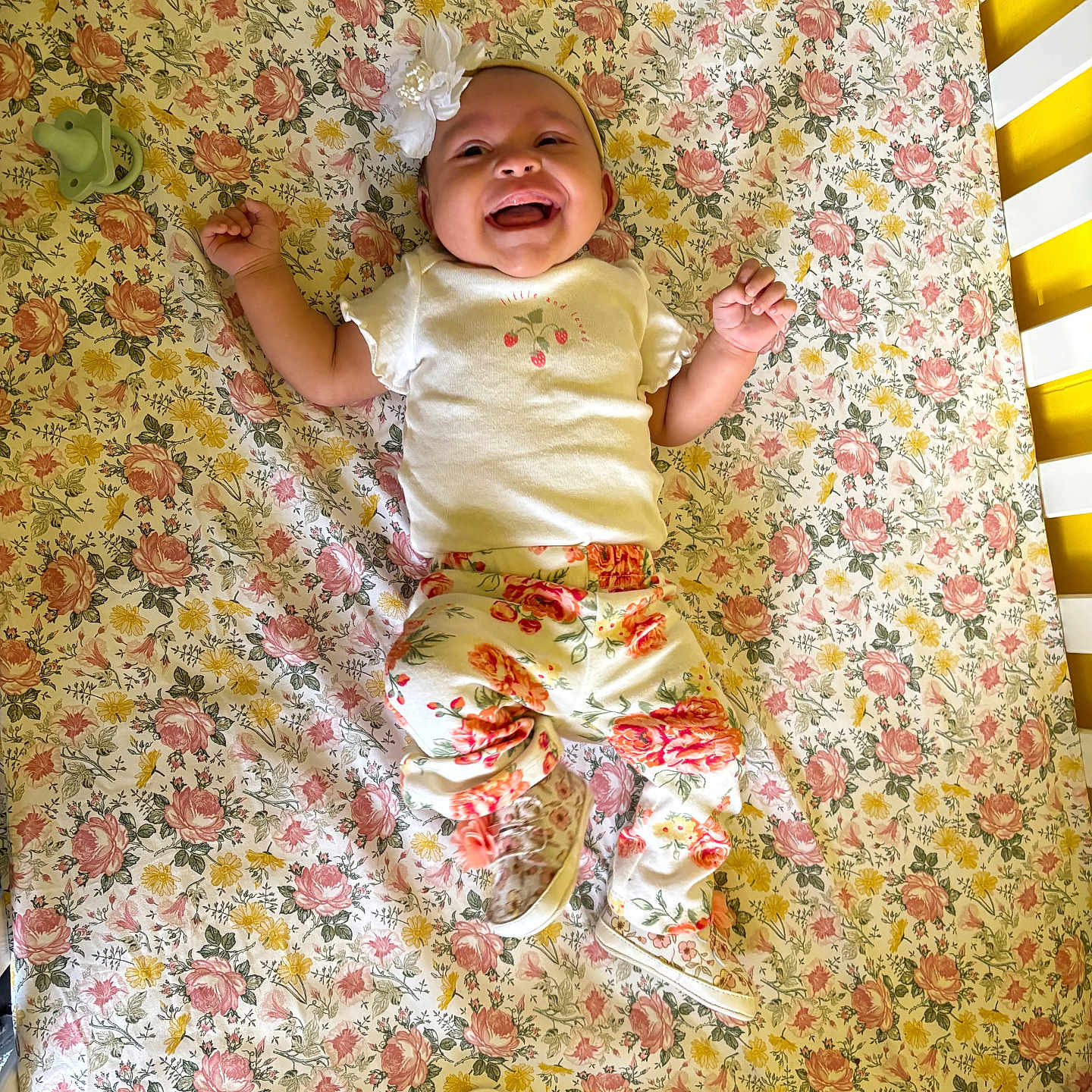 Octavia is registered to the contest to win money with this photo: baby, baby_clothes, bed, child, crib, cute, fabric, floral_pattern, flower, footwear, happy, headband, infant, lying_down, nursery, pacifier, patterned_pants, smiling, toy, white_top