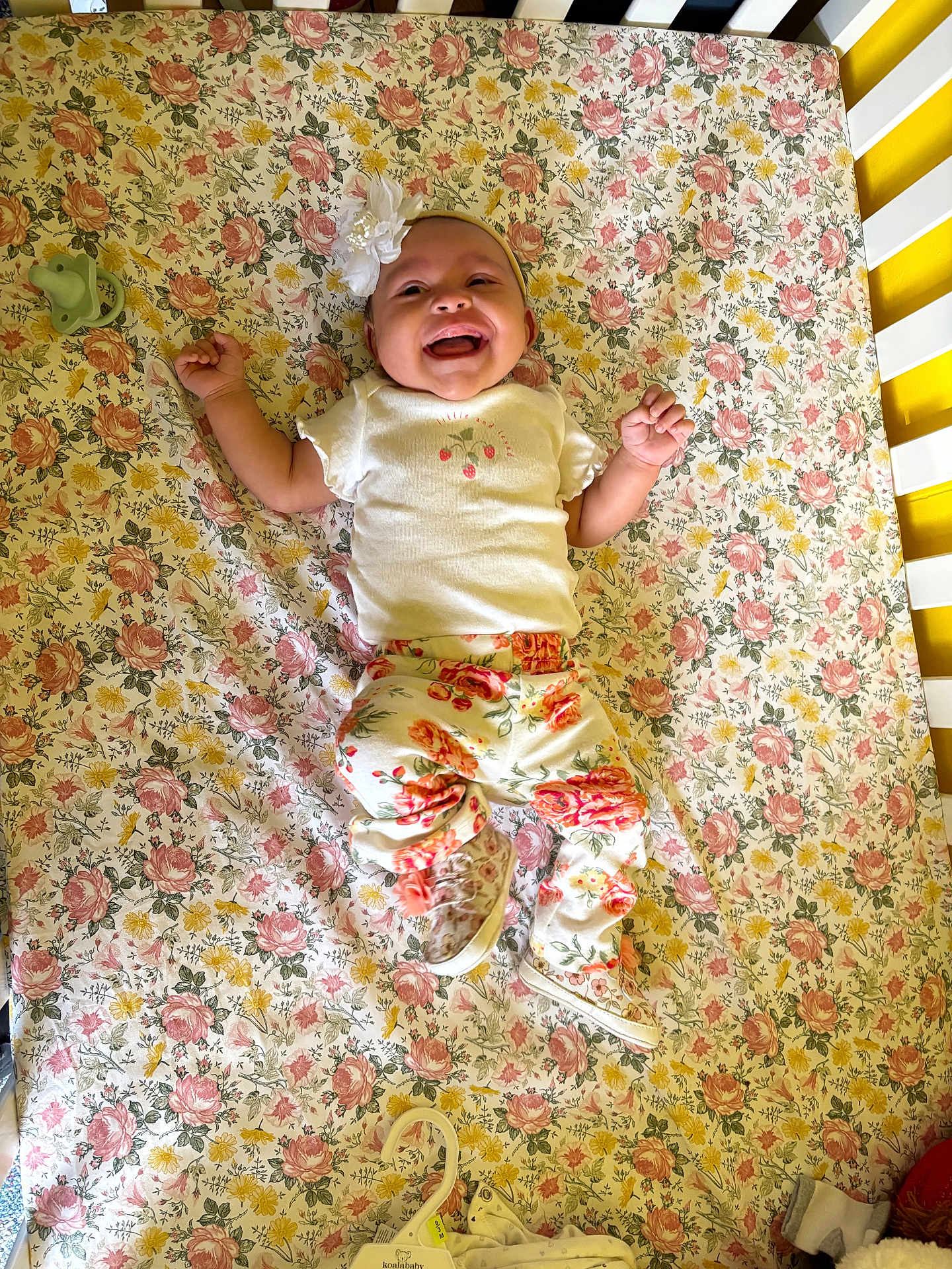 Octavia is registered to the contest to win money with this photo: baby, smiling, crib, floral_pattern, headband, pacifier, baby_clothes, infant, lying_down, happy, white_top, patterned_pants, nursery, cute, child, flower, fabric, footwear, toy, bed