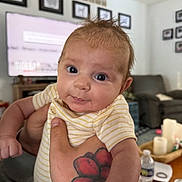Magnolia is registered to the contest to win money with this photo: baby, infant, newborn, hand, tattoo, onesie, striped_clothing, wide_eyes, curious_expression, living_room, couch, television, coffee_table, water_bottle, candles, portrait, person, indoor, furniture, family_room