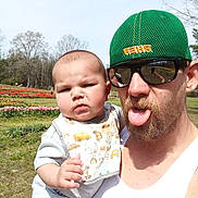 Jackson is registered to the contest to win money with this photo: baby, beard, bib, cap, casual_clothing, child, father, flowers, grass, holding, man, outdoor, portrait, selfie_reflection, sky, sunglasses, sunlight, tongue_out, trees, tulip_field